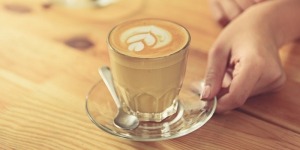 close up of female hands holding cup of delicious latte with tulip art on wooden worktop. Woman standing at bar counter in cafe